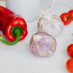 Fresh garlic placed among ripe red cherry tomatoes and peppers on white table near white ceramic cup and yellow lemon